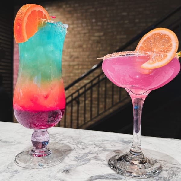 Two vibrant cocktails, one layered blue, red, and purple, the other pink, on a marble bar.