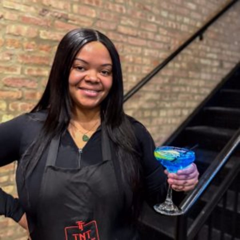 A smiling TNT Rooftop Restaurant staff member holds a blue cocktail with a lime garnish.