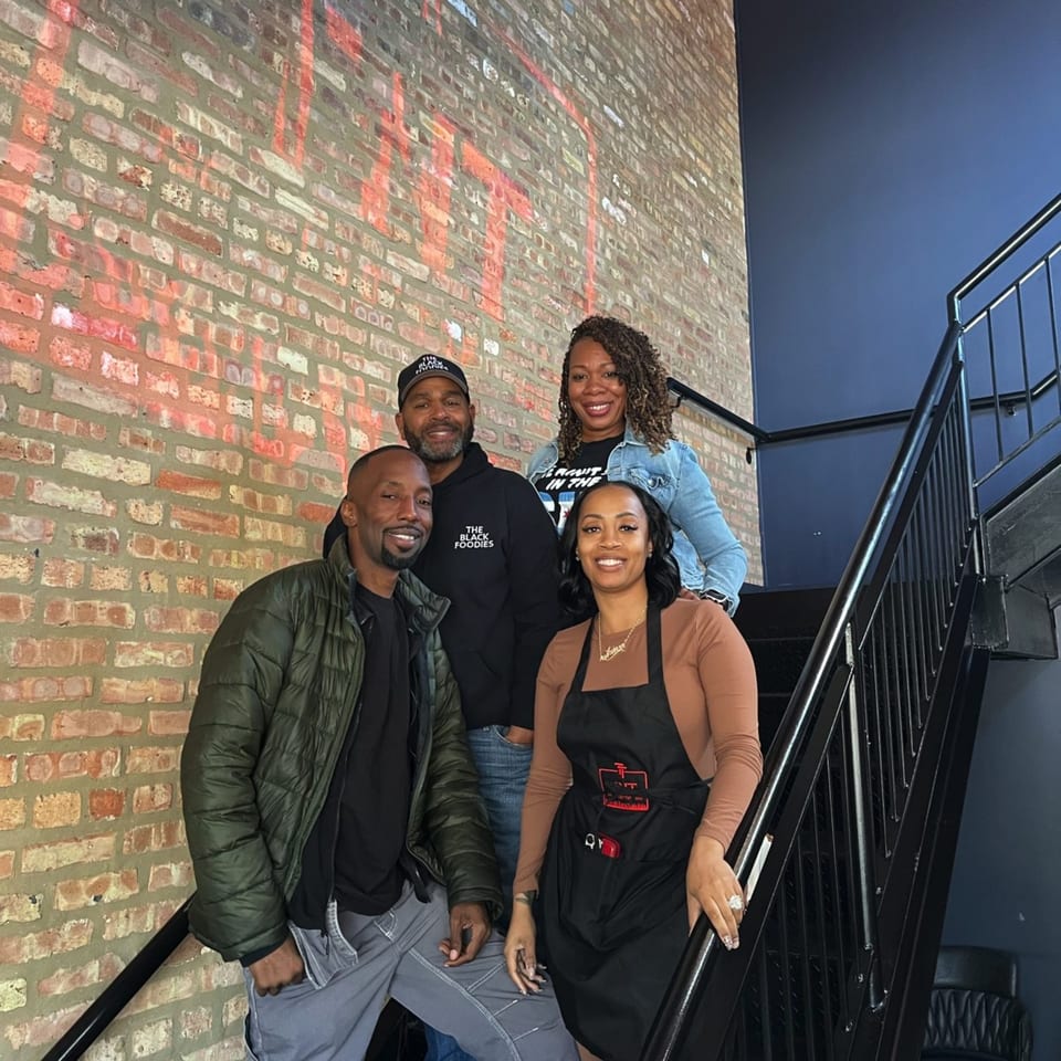Four smiling TNT Rooftop Restaurant staff members pose on a black metal staircase against a brick wall.