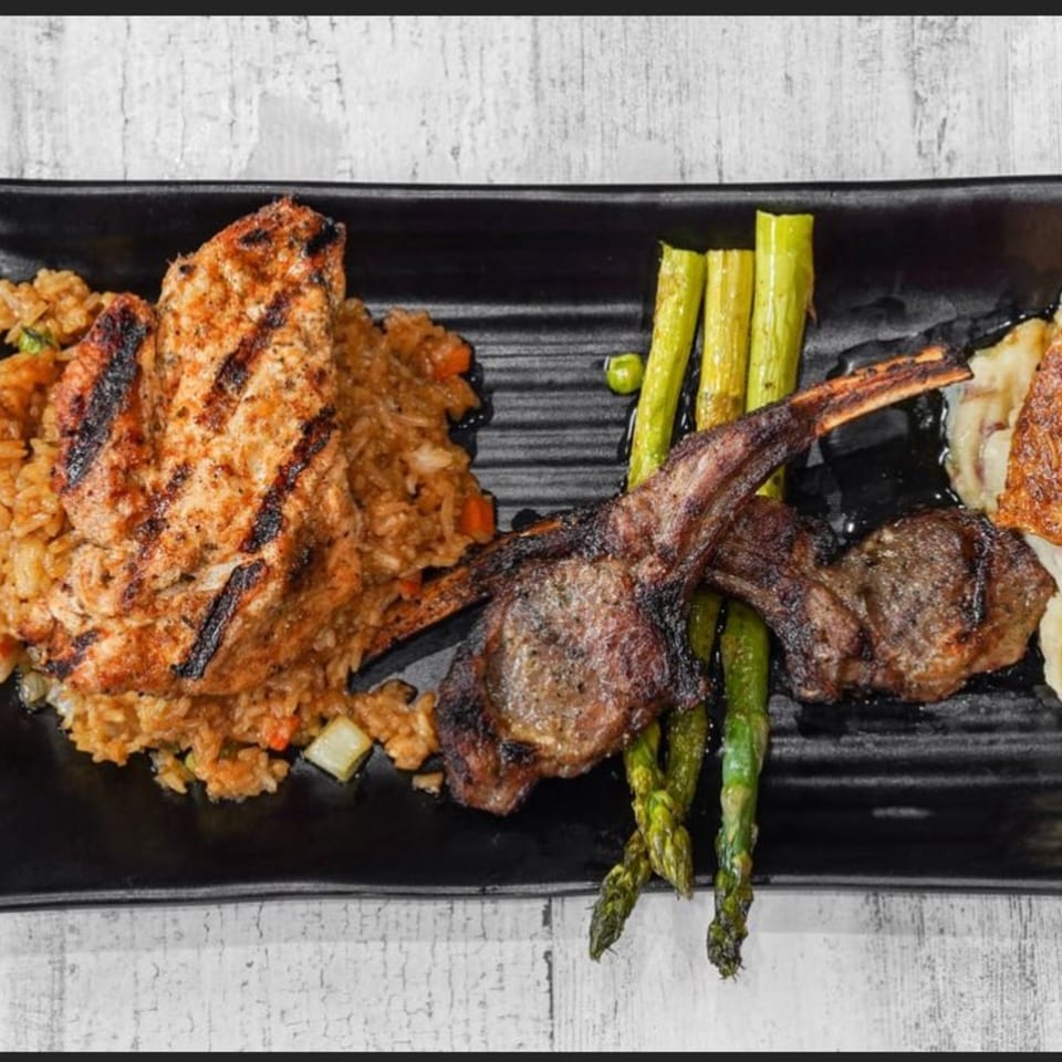 Grilled chicken and lamb chops with rice and asparagus on a black rectangular plate.